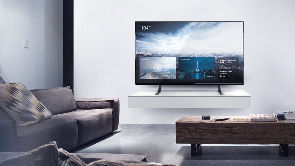 W nowoczesnym salonie znajduje się duży telewizor zamontowany na ścianie nad białą półką. Przed telewizorem stoi drewniany stolik, a obok znajduje się szara sofa z poduszkami.