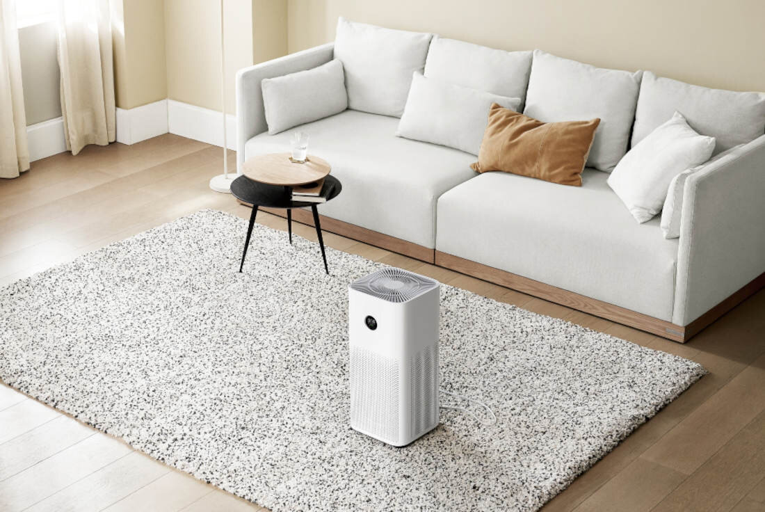 W salonie na jasnym dywanie stoi biały oczyszczacz powietrza. W tle znajduje się jasna sofa z kilkoma poduszkami oraz mały stolik kawowy.
