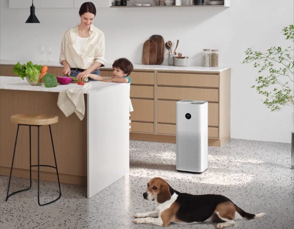 W kuchni stoi biały oczyszczacz powietrza z okrągłym wyświetlaczem. Na podłodze leży pies, a przy blacie kuchennym znajdują się dwie osoby przygotowujące jedzenie.