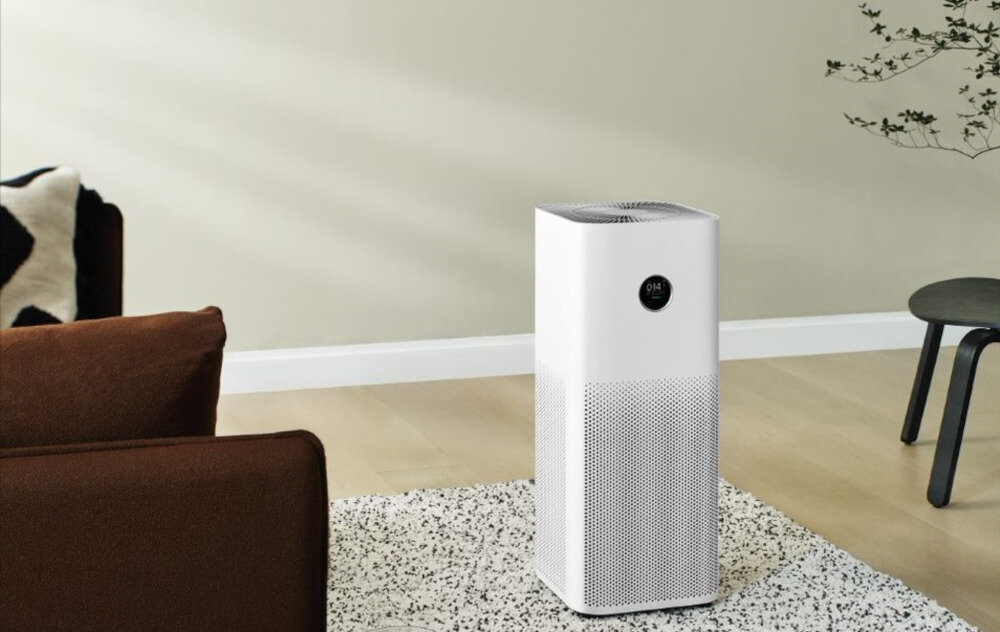 Na zdjęciu znajduje się biały oczyszczacz powietrza o prostokątnym kształcie, umieszczony na jasnej podłodze obok brązowej sofy i małego stolika. Na oczyszczaczu widoczny jest okrągły wyświetlacz z cyframi \'014\'.