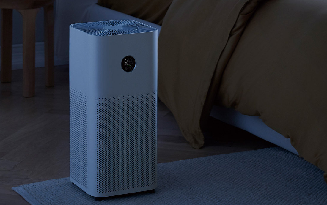 Na obrazie znajduje się biały oczyszczacz powietrza z okrągłym wyświetlaczem pokazującym wartość \'014\'. Urządzenie stoi na podłodze obok łóżka z brązową pościelą.