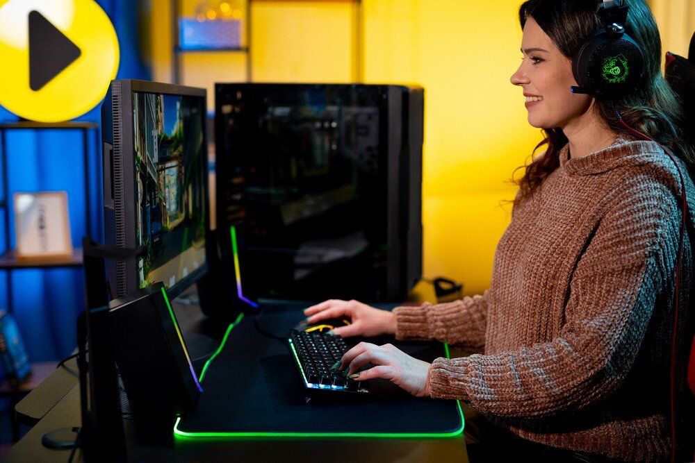 Komputer MAD DOG GeForce RTX5070Ti PurePC Edition 4 Widok od boku, kobieta w słuchawkach gamingowych siedząca przy biurku grająca w grę, w tle komputer wygląd opis