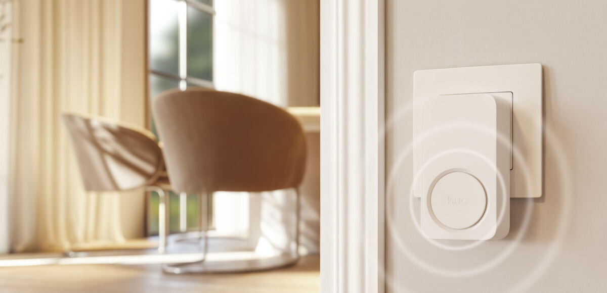 Dzwonek PHILIPS HUE, dom, części, niezależne, pokoój, Minimalistyczny sygnalizator Hue Secure umieszczony w gniazdku elektrycznym na tle jasnego, nowoczesnego wnętrza z rozmytym fotelem w tle.