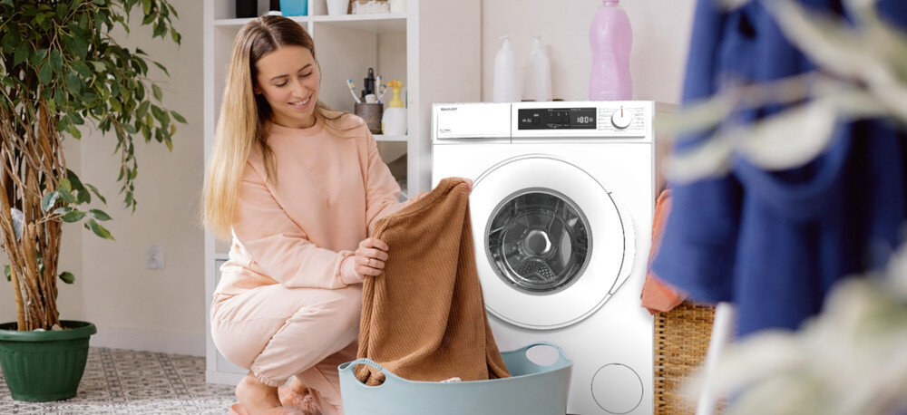 PRALKA SHARP ES-NFA6101WC-PL Osoba wkłada ubrania do bębna pralki ustawionej w domowym wnętrzu. Widoczny jest kosz na pranie i półki z detergentami pranie wełny delikatne tkaniny łagodny cykl ochrona ubrań pielęgnacja materiałów