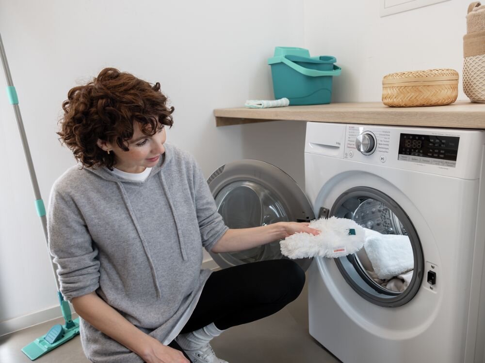 Zmiotka do kurzu LEIFHEIT Superduster Kobieta w szarej bluzie i czarnych legginsach wkłada białą końcówkę miotełki do kurzu Leifheit do otwartych drzwi pralki w jasnej pralni. Obok pralki znajduje się plastikowe wiaderko w turkusowym kolorze i pojemnik z naturalnego materiału. W tle widoczne są środki czystości i mop