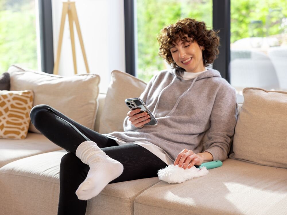 Zmiotka do kurzu LEIFHEIT Superduster Kobieta odpoczywa na beżowej kanapie w dobrze oświetlonym salonie, ubrana w szarą bluzę i czarne legginsy, z białą miotełką do kurzu Leifheit w dłoni i smartfonem w drugiej ręce. Na kanapie leżą jasne poduszki, a za oknem widoczna jest zieleń ogrodu