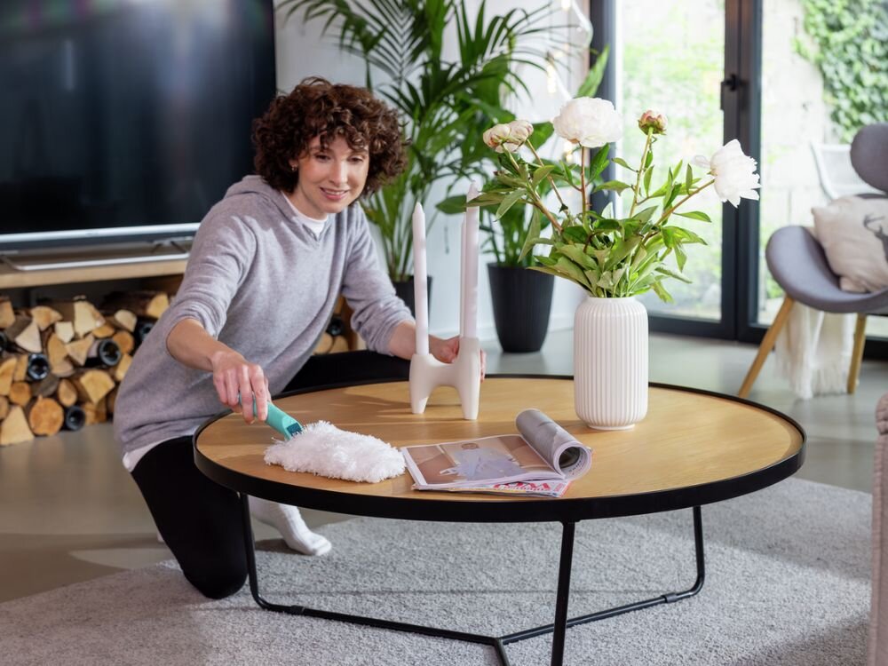 Zmiotka do kurzu LEIFHEIT Superduster Kobieta w szarej bluzie i czarnych legginsach klęczy na dywanie w nowoczesnym salonie i wyciera okrągły, jasny stolik na czarnej ramie białą miotełką do kurzu z turkusową rączką. Na stoliku stoją świecznik z trzema świecami, wazon z białymi kwiatami oraz gazeta. W tle widać telewizor, drewno kominkowe i rośliny doniczkowe.
