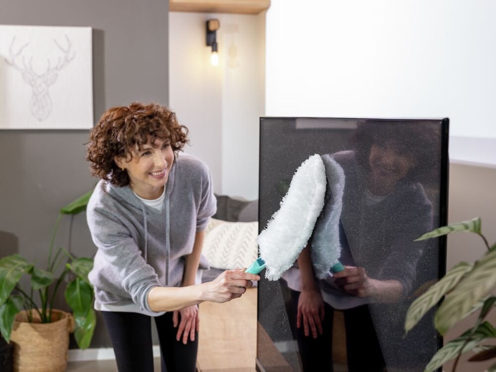 Zmiotka do kurzu LEIFHEIT Superduster XL Kobieta ubrana w szarą bluzę i czarne legginsy czyści poziome żaluzje dużym, białym pióropuszem do kurzu Leifheit z turkusową rączką w nowoczesnym, jasnym wnętrzu z dużymi oknami, przez które widać ogród.