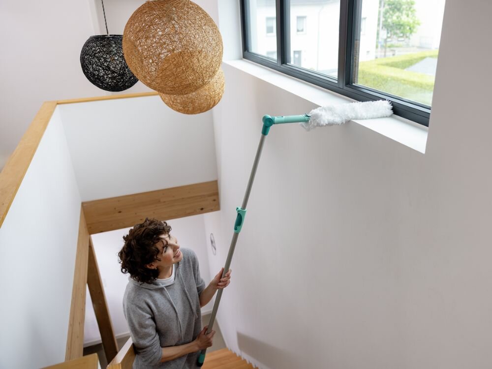 Zmiotka do kurzu LEIFHEIT Superduster XL Kobieta odpoczywa na beżowej kanapie w dobrze oświetlonym salonie, ubrana w szarą bluzę i czarne legginsy, z białą miotełką do kurzu Leifheit w dłoni i smartfonem w drugiej ręce. Na kanapie leżą jasne poduszki, a za oknem widoczna jest zieleń ogrodu.