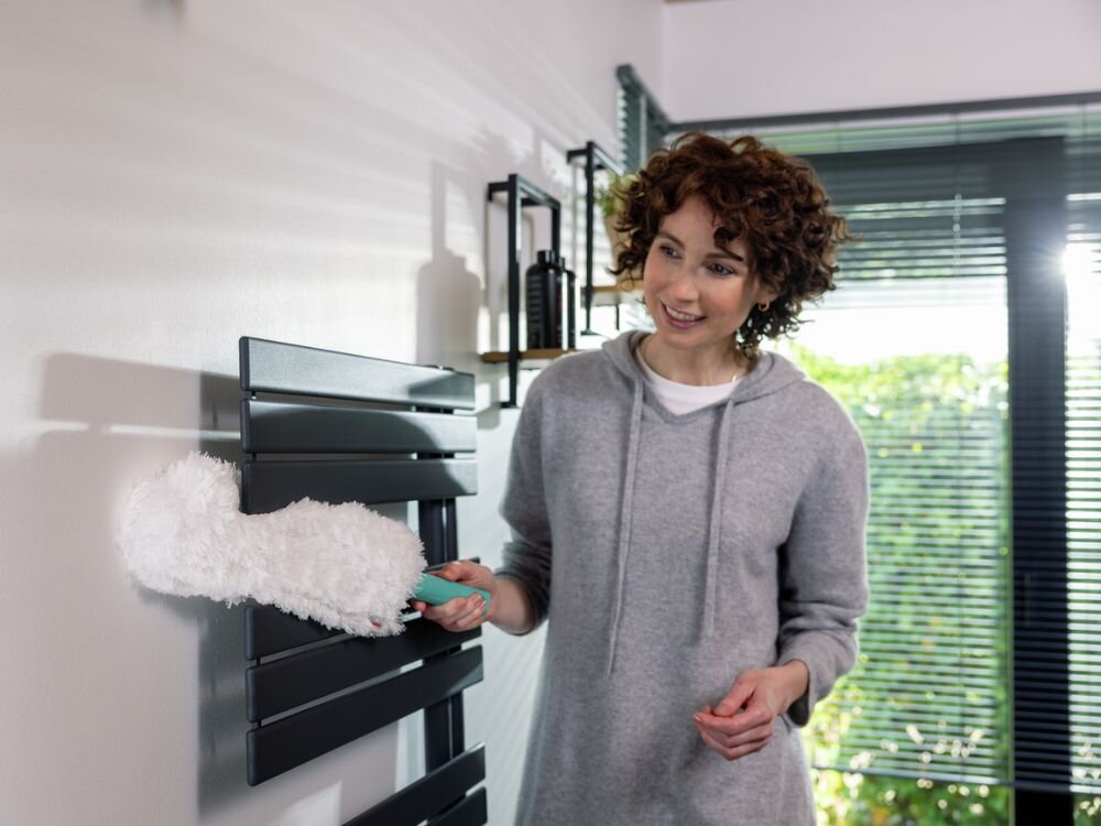 Zmiotka do kurzu LEIFHEIT Superduster XL Kobieta w szarej bluzie i czarnych legginsach klęczy na dywanie w nowoczesnym salonie i wyciera okrągły, jasny stolik na czarnej ramie białą miotełką do kurzu z turkusową rączką. Na stoliku stoją świecznik z trzema świecami, wazon z białymi kwiatami oraz gazeta. W tle widać telewizor, drewno kominkowe i rośliny doniczkowe.