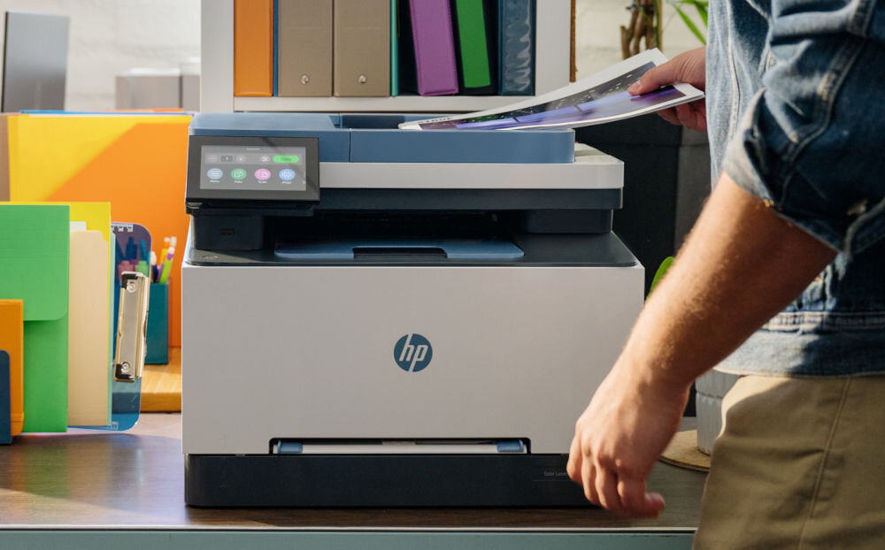 Na biurku stoi urządzenie wielofunkcyjne z logo HP, obok którego znajduje się osoba wkładająca dokument do podajnika. W tle widoczne są kolorowe segregatory i przybory biurowe.