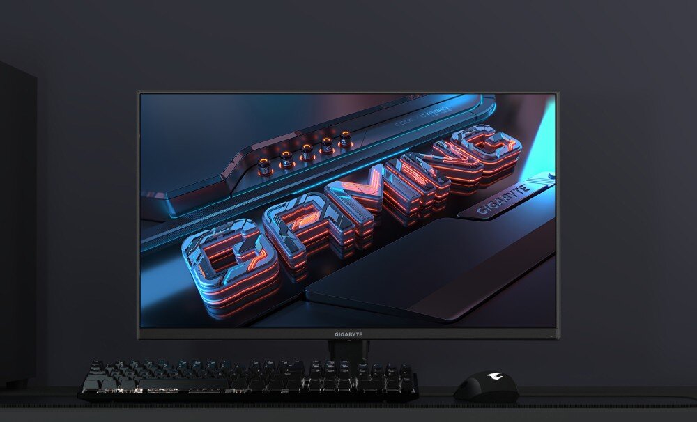 Monitor GIGABYTE M32UP Urządzenie stojące na biurku z klawiaturą i myszką wyświetlające trójwymiarowy napis 'GAMING', opis