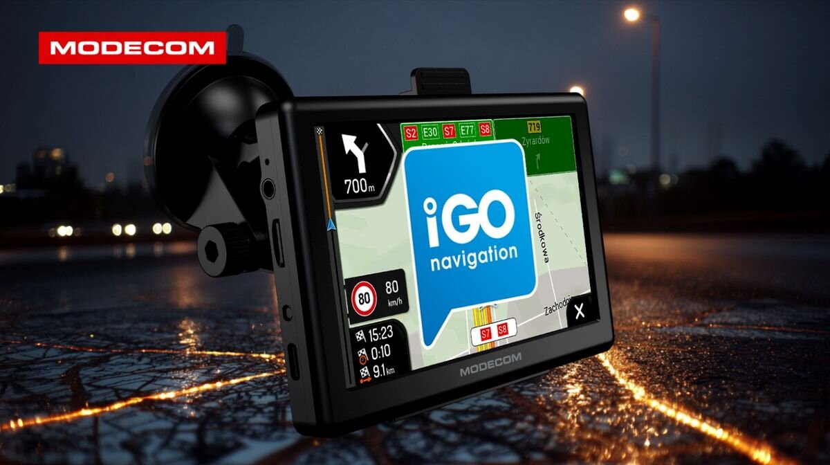 Urządzenie nawigacyjne z logo \'MODECOM\' i ekranem wyświetlającym mapę oraz logo \'iGO navigation\'. W tle widoczna jest rozmyta sceneria nocna z oświetleniem ulicznym.