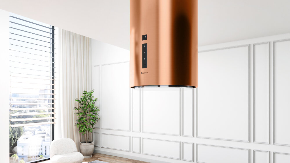 Okap GLOBALO Heweno Isola 39.2 Miedziany mat Na zdjęciu przedstawiono przestronną kuchnię w minimalistycznym stylu, z centralnie umieszczoną wyspą wykonaną z jasnego, marmurowego kamienia. Nad wyspą zawieszony jest efektowny okap cylindryczny, który stanowi designerski akcent wnętrza. W tle widać wysokie drewniane zabudowy piekarnik w zabudowie, a całość uzupełnia eleganckie krzesło i zielona roślina w doniczce szerokość 39 cm timer czas Głośność 62 db