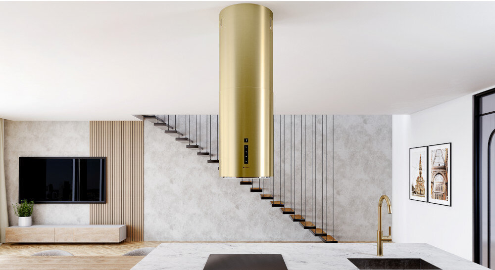 Okap GLOBALO Heweno Isola 39.2 Złoty mat Na zdjęciu widoczny jest matowy złoty okap kuchenny o cylindrycznym kształcie, zawieszony centralnie nad wyspą, w eleganckim i jasnym wnętrzu z widocznymi schodami, telewizorem i obrazami na ścianie. Całość podkreśla luksusowy charakter kuchni oraz minimalistyczny design