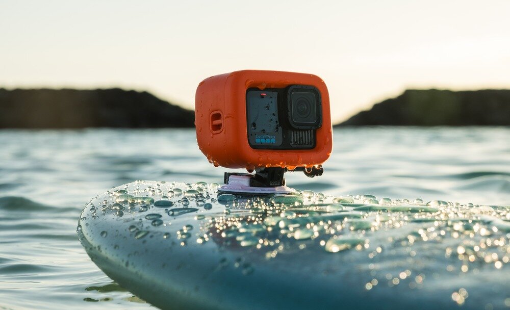 Kamera sportowa w pomarańczowej obudowie zamontowana na desce surfingowej na wodzie. W tle widać rozmyty krajobraz z ciemnymi skałami i jasnym niebem.