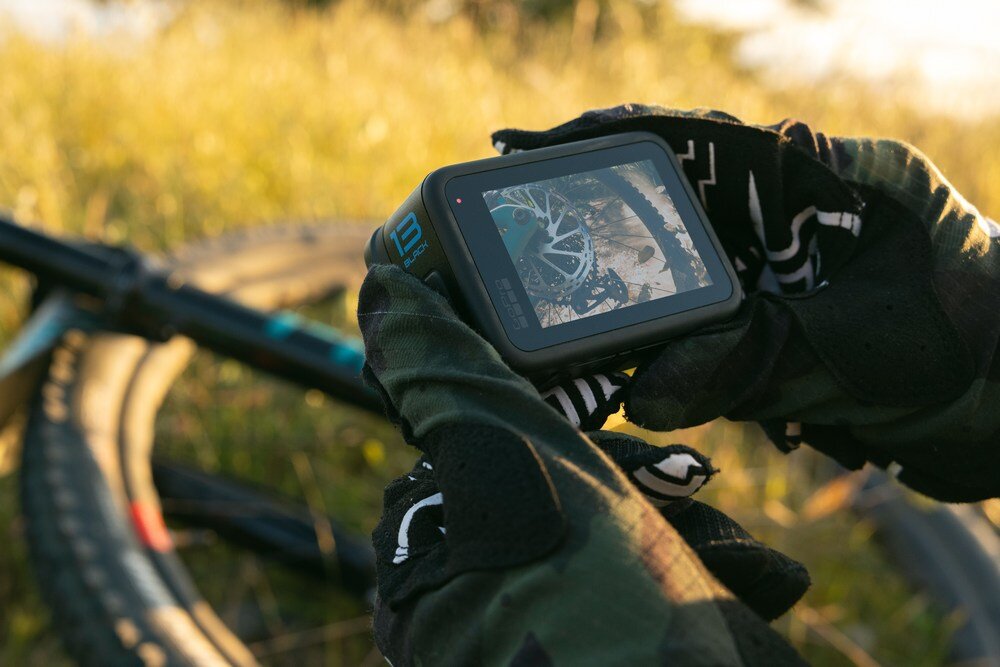 Osoba w rękawiczkach trzyma kamerę sportową, na której ekranie widać obraz roweru. W tle znajduje się trawa oświetlona słońcem.