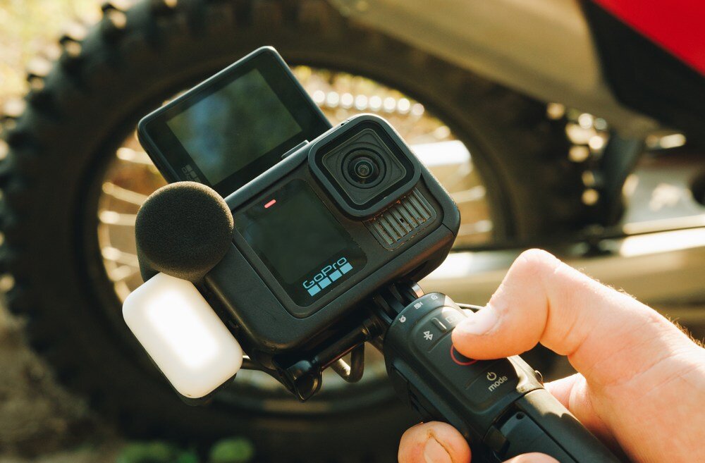 Osoba trzyma kamerę sportową z logo \'gopro\' na uchwycie, z widocznym ekranem i mikrofonem. W tle znajduje się koło motocykla.