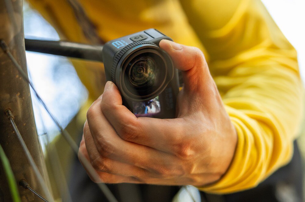 Osoba trzyma kamerę sportową z widocznym obiektywem i logo \'m\' na obudowie. W tle widać fragment roweru i drzewo.