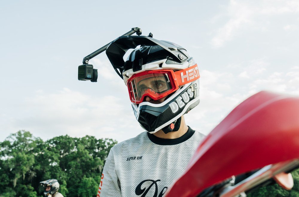 Osoba w kasku motocyklowym z zamontowaną kamerą sportową na szczycie. Na kasku widoczne są napisy \'HEAT\' i \'DEUS\'.