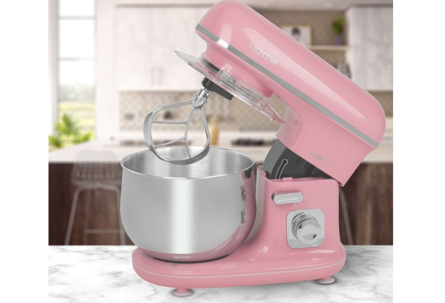 Różowy robot kuchenny z metalową misą i mieszadłem, ustawiony na blacie kuchennym. W tle widoczna jest kuchnia z szafkami i oknem.