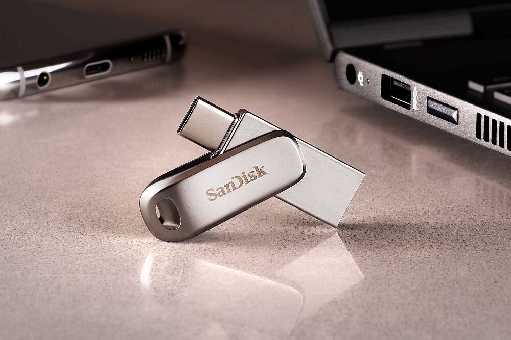 Na obrazie widoczny jest srebrny pendrive z logo SanDisk, umieszczony na powierzchni obok laptopa. Pendrive ma dwa złącza: USB-C i USB 3.0.