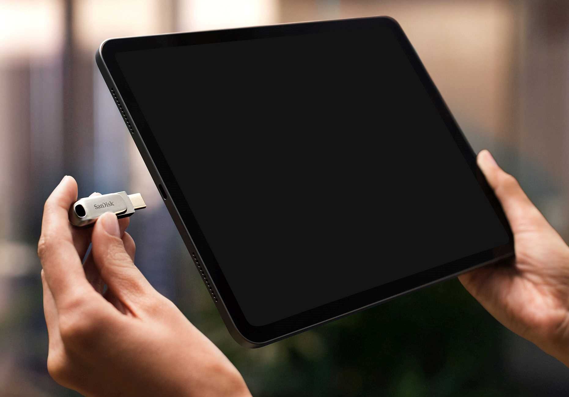 Osoba trzyma tablet oraz pendrive z logo SanDisk, który jest w trakcie podłączania do urządzenia.