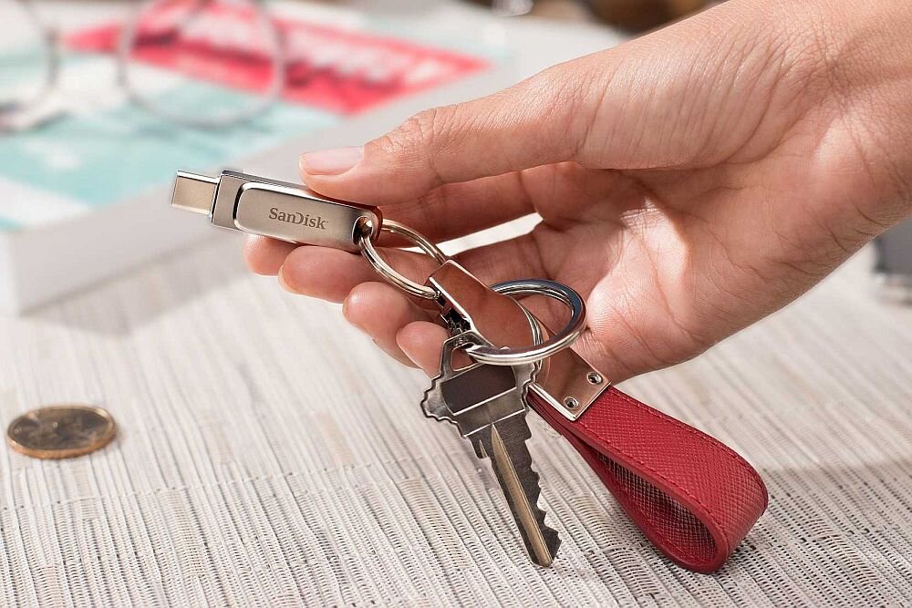 Osoba trzyma brelok z kluczami i pendrive\'em marki SanDisk. Pendrive ma metaliczną obudowę i jest podłączony do kółka na klucze.