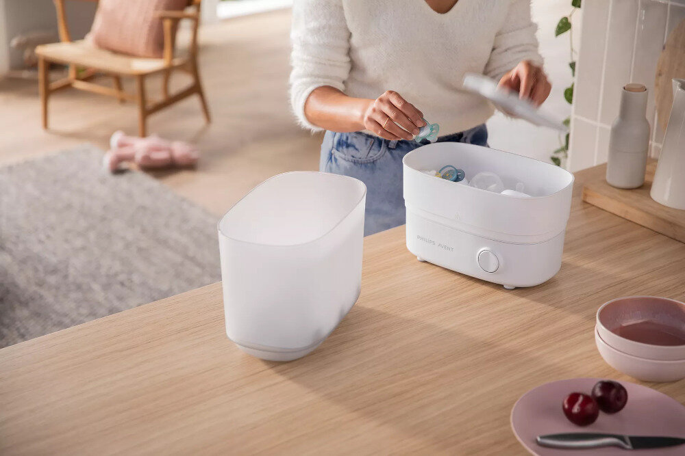 Na drewnianym blacie znajduje się elektryczny sterylizator parowy z otwartą pokrywą, w którym umieszczone są smoczki. Obok stoi osoba, która trzyma pokrywkę, a na blacie widoczne są również miski i owoce.