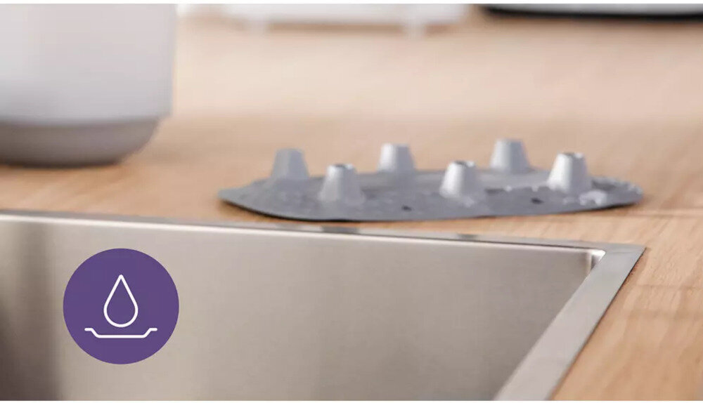 Na drewnianym blacie kuchennym znajduje się metalowy zlew oraz szara, plastikowa tacka z wypustkami. W lewym dolnym rogu widoczna jest fioletowa ikona z symbolem kropli wody.