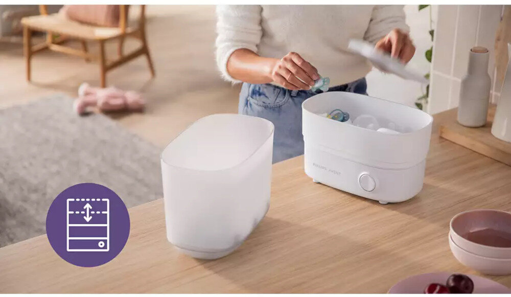 Na blacie kuchennym znajduje się biały elektryczny sterylizator parowy z logo Philips Avent, obok którego stoi osoba wkładająca do niego smoczek. Obok urządzenia leży przezroczysta pokrywa, a na blacie widoczne są miski z owocami.