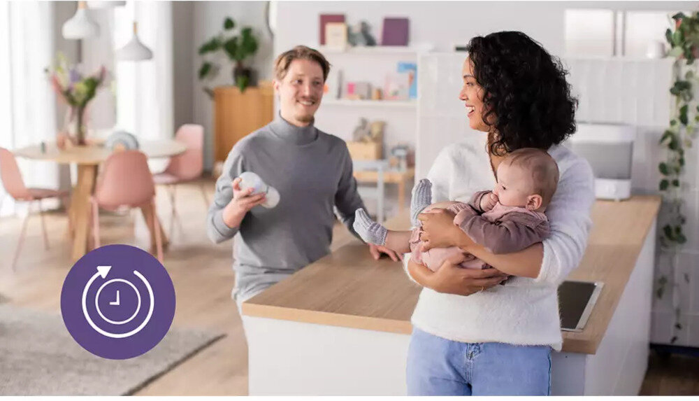W jasnym wnętrzu kuchni stoi kobieta trzymająca dziecko, a obok niej mężczyzna z pluszową zabawką. Na blacie widoczny jest elektryczny sterylizator parowy, a w rogu zdjęcia znajduje się ikona zegara na fioletowym tle.