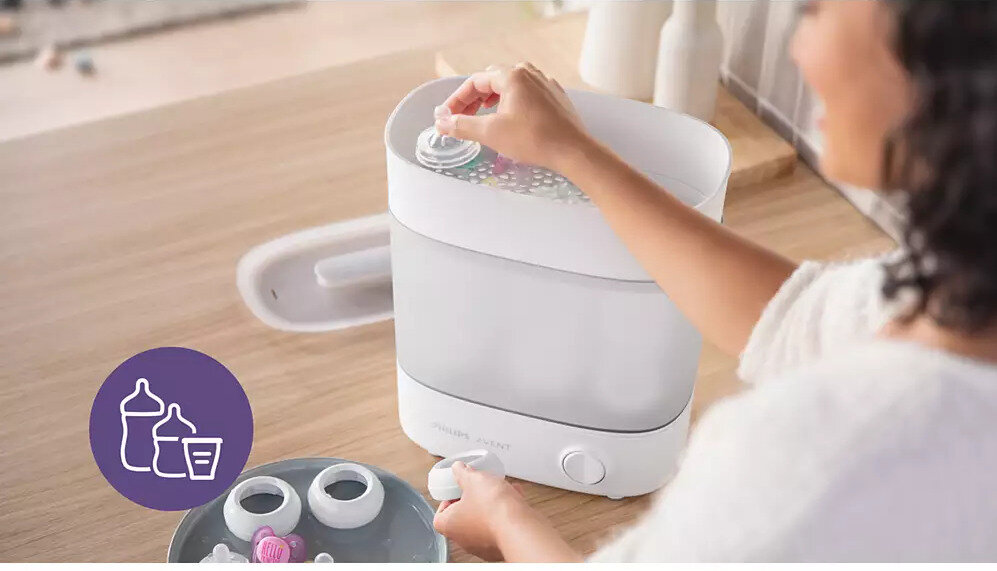 Osoba obsługuje elektryczny sterylizator parowy na drewnianym blacie. Obok znajduje się okrągła ikona z białymi konturami butelek na fioletowym tle.