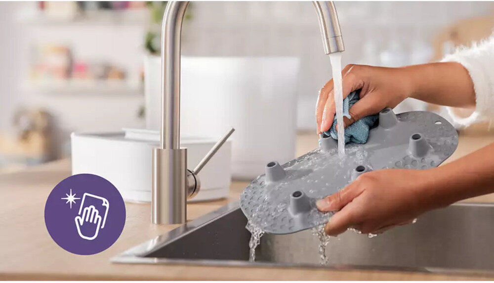 Osoba myje szarą plastikową część pod bieżącą wodą w zlewie. W lewym dolnym rogu znajduje się fioletowa ikona z symbolem dłoni i błyskiem.