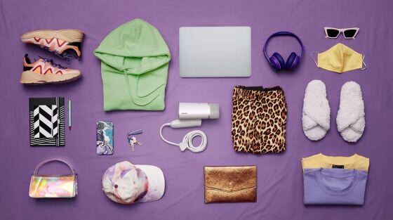 Na fioletowym tle leżą różne przedmioty, w tym zielona bluza, laptop, słuchawki, parownica do ubrań z napisem \'PHILIPS\', spodenki w panterkę, kapcie, maska, okulary, torebka, czapka, buty sportowe, notatnik, długopis, telefon i portfel. Wszystkie przedmioty są ułożone w estetyczny sposób.