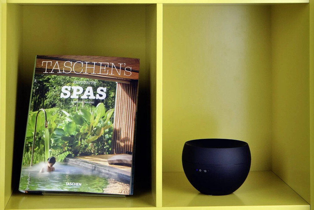 Na żółtej półce znajduje się książka z napisem \'TASCHEN\'s FAVORITE SPAS\' oraz czarny aromatyzer o gładkiej powierzchni z kilkoma małymi diodami.