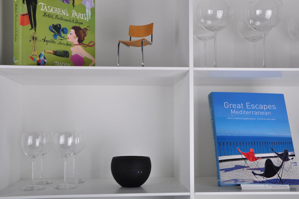Na półce znajduje się czarny aromatyzer o zaokrąglonym kształcie. Obok niego stoją trzy kieliszki oraz książka z niebieską okładką zatytułowana \'Great Escapes Mediterranean\'.