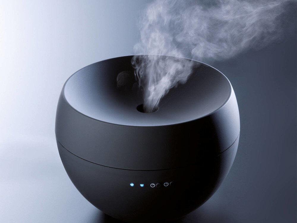 Czarny aromatyzer o zaokrąglonym kształcie z otworem na górze, z którego unosi się para. Na urządzeniu widoczne są trzy niebieskie diody oraz przyciski z symbolami zasilania i ustawień.