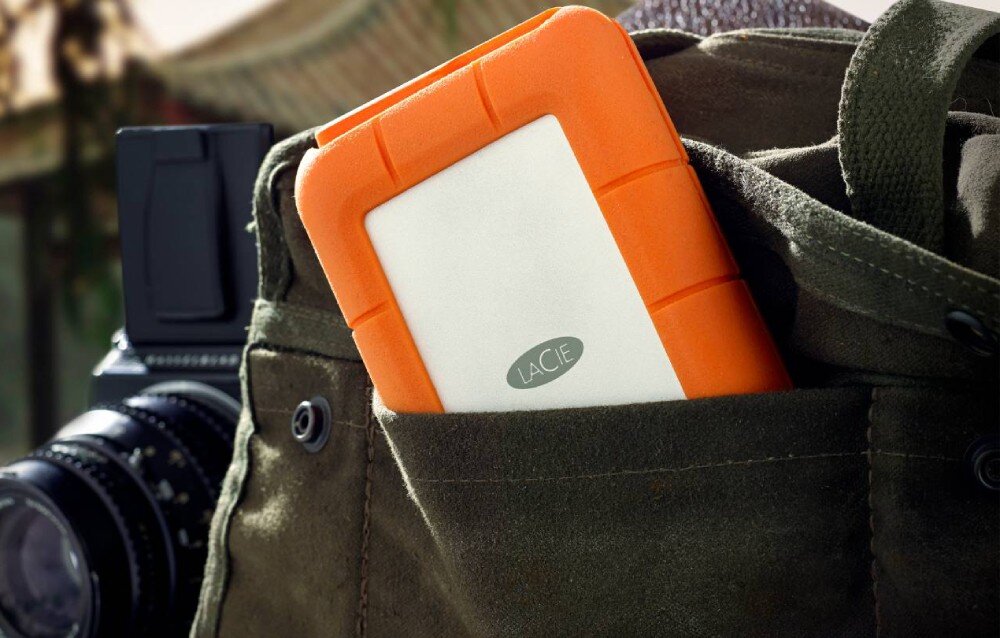 Dysk LACIE Rugged SSD4 Pomarańczowo-srebrny dysk przenośny włożony do kieszeni torby z materiału, obok aparatu fotograficznego z widocznym obiektywem, opis