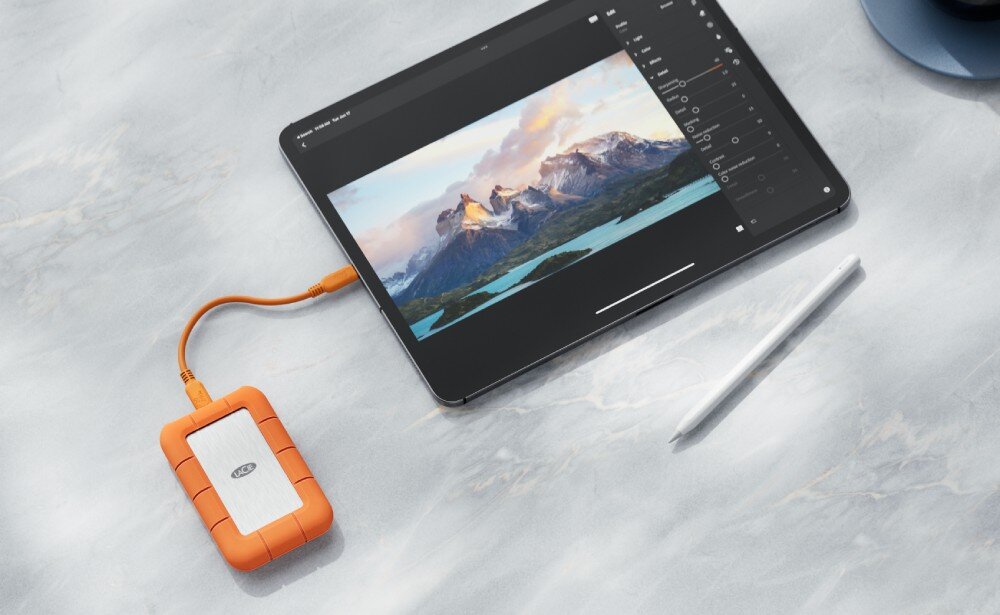 Dysk LACIE Rugged SSD4 Pomarańczowo-srebrny dysk przenośny połączony pomarańczowym kablem z tabletem, na którego ekranie wyświetlane jest zdjęcie górskiego krajobrazu, obok leży biały rysik, cechy, zalety, interfejs