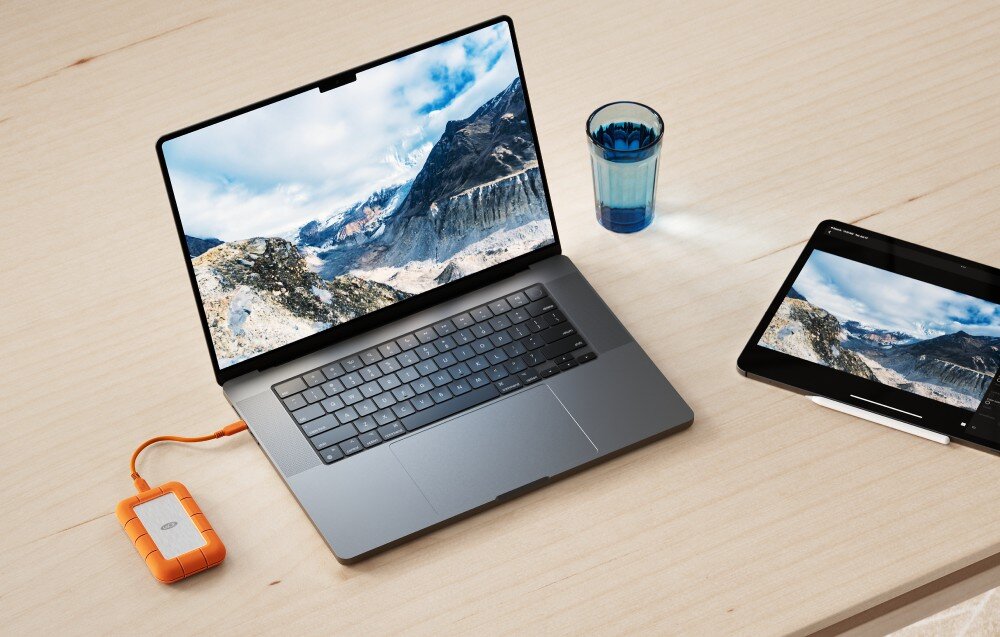 Dysk LACIE Rugged SSD4 Pomarańczowo-srebrny dysk przenośny podłączony do laptopa z wyświetlonym zdjęciem gór, obok stoi szklanka z wodą i tablet z tym samym obrazem, konstrukcja, wytrzymałość, szczelność IP54