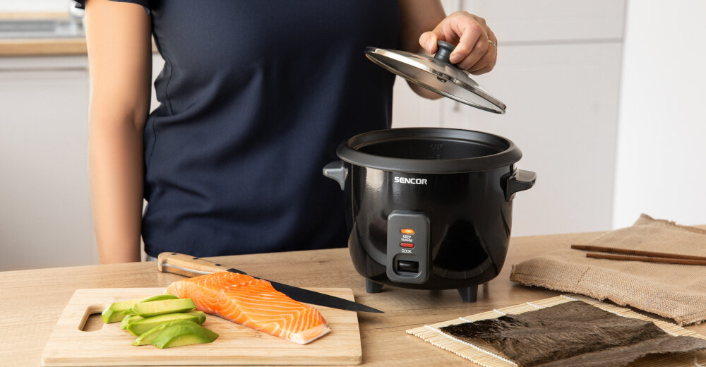 Osoba trzyma pokrywkę ryżowaru marki SENCOR, który stoi na blacie obok deski z pokrojonym łososiem i awokado. Obok leży nóż oraz arkusze nori.
