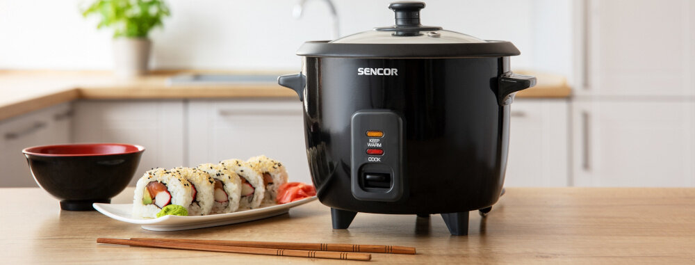 Czarny ryżowar z logo SENCOR stoi na blacie kuchennym obok talerza z sushi i pałeczek. W tle widoczna jest kuchnia z rośliną w doniczce.