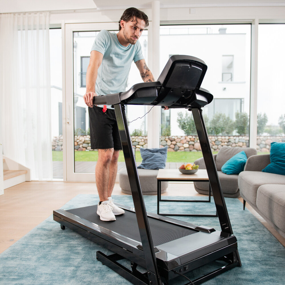 Bieżnia elektryczna KETTLER Track 100 1287W 16 km-h dom pokój salon osoba opiera się o bieżnię w tle okno za oknem ogród regularny trening bez wychodzenia z domu zakres prędkości 1–16 km/h pas biegowy o wymiarach 125 × 42 cm swoboda ruchu i komfort składana konstrukcja wbudowane programy treningowe