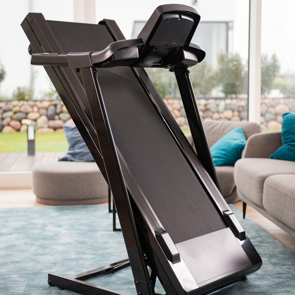 Bieżnia elektryczna KETTLER Track 100 1287W 16 km-h dom pokój salon złożona bieżnia w salonie w tle okno za oknem ogród funkcja składania kółka transportowe mechanizm Soft Drop pas biegowy opuszcza się powoli i bezpiecznie czas montażu około 15 minut wymiary po złożeniu 68 x 131 x 94 cm