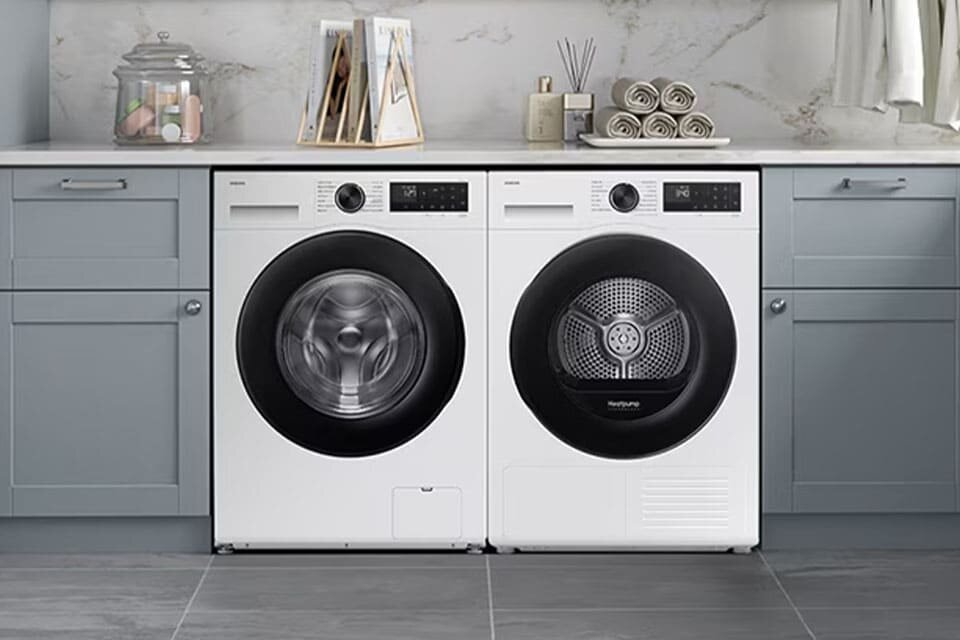 Pralka AI EcoBubble™ 10kg WW10FG5U34AEEO w zabudowie Biała pralka i suszarka Samsung obok siebie pod blatem w jasnej pralni z szarymi szafkami i ręcznikami - dostępna w Media Expert