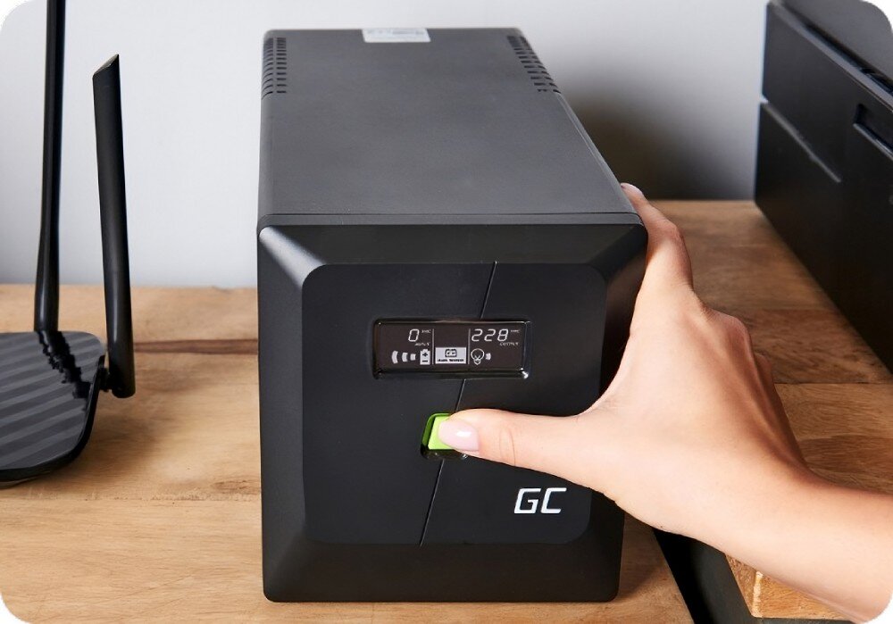 Zasilacz UPS GREENCELL UPSLM1200 Ręka naciska zielony przycisk na czarnym urządzeniu z wyświetlaczem pokazującym napięcie i ikonami stanu, stojącym na drewnianym blacie obok routera z antenami, opis
