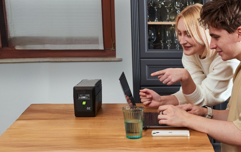 Zasilacz UPS GREENCELL UPSLM480 Dwie osoby siedzą przy drewnianym stole z laptopem, obok którego stoi czarny zasilacz z wyświetlaczem i zielonym przyciskiem, na stole leży też szklanka z wodą i telefon, opis