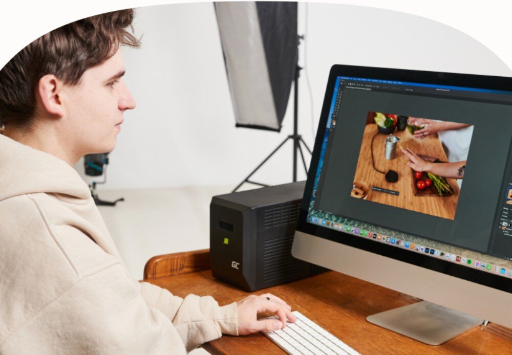 Zasilacz UPS GREENCELL UPSLM480 Mężczyzna siedzi przy biurku i pracuje na komputerze stacjonarnym, obok monitora stoi czarny zasilacz z widocznym wyświetlaczem i zielonym przyciskiem, zastosowanie, działanie