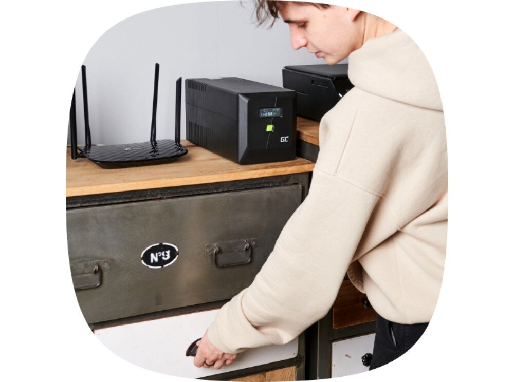 Zasilacz UPS GREENCELL UPSLM480 Mężczyzna w jasnej bluzie sięga do szuflady, a na blacie obok routera stoi czarny zasilacz z wyświetlaczem LCD i zielonym przyciskiem, konstrukcja, budowa,